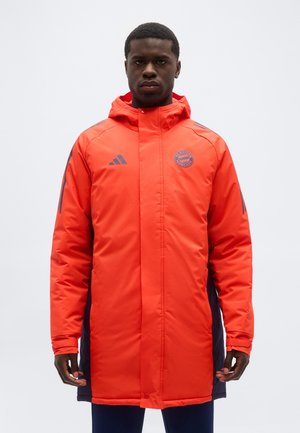 Mann trägt eine lange, leuchtend orangefarbene, gepolsterte FC Bayern München Jacke mit Kapuze, steht vor einem schlichten weißen Hintergrund.