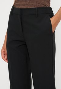 Gros plan d'une personne portant un pantalon noir sur mesure avec les mains près des poches et un haut marron texturé rentré.