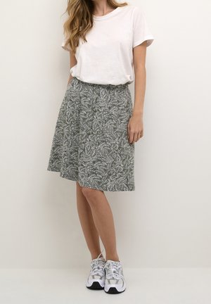 Kvinde iført hvid t-shirt, knælængde sort-hvidt blomsterprint nederdel og hvide snørelørere sneakers, stående foran en ensfarvet væg.