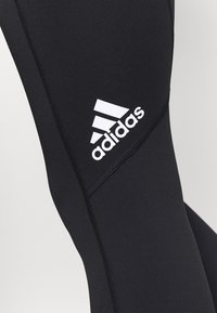 Leggings noirs en matériau extensible, présentant un logo Adidas blanc et des détails de couture contrastants le long des coutures.