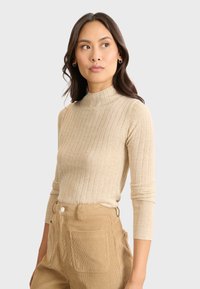 Beige geripptes Rollkragenoberteil mit langen Ärmeln, aus leichtem Strickstoff, weicher Textur und figurbetontem Schnitt. Kombiniert mit hellbraunen Cordhosen.