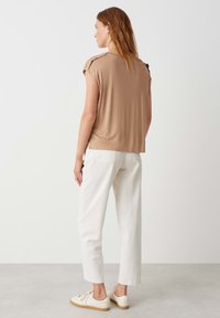 Tan korte mouwen top met een relaxed fit, gecombineerd met witte recht afgesneden broeken en beige sneakers op een lichte achtergrond.