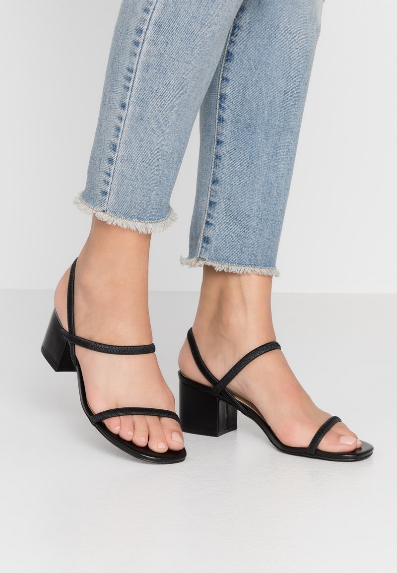 Sandales noires à talons avec deux fines sangles sur les orteils, un talon carré et une texture lisse. Associées à un jean bleu avec un ourlet effiloché.