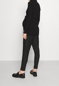 Schwangere Person, die ein schwarzes Langarmoberteil, gestreifte schwarze Hosen und schwarze Slipper trägt, steht vor einem schlichten weißen Hintergrund.
