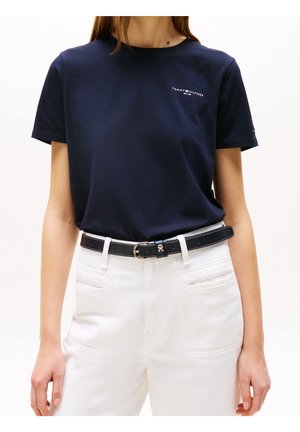 Kobieta nosząca granatową koszulkę Tommy Hilfiger włożoną do białych spodni z czarnym paskiem, stojąca na białym tle.