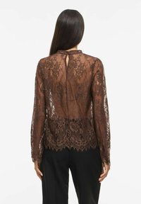 Femme aux longs cheveux foncés portant un chemisier en dentelle marron transparent à motifs floraux et un pantalon noir, vue de dos sur fond blanc.