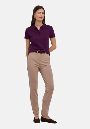 Vrouw stond met handen achter de rug, gekleed in een paarse polo met korte mouwen, beige strakke broek, een zwarte riem en zwarte platte schoenen.