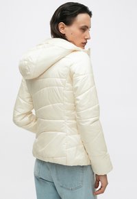 Giacca puffy crema con cappuccio, caratterizzata da dettagli con cuciture orizzontali, una superficie liscia e un design aderente, abbinata a jeans azzurri chiari.