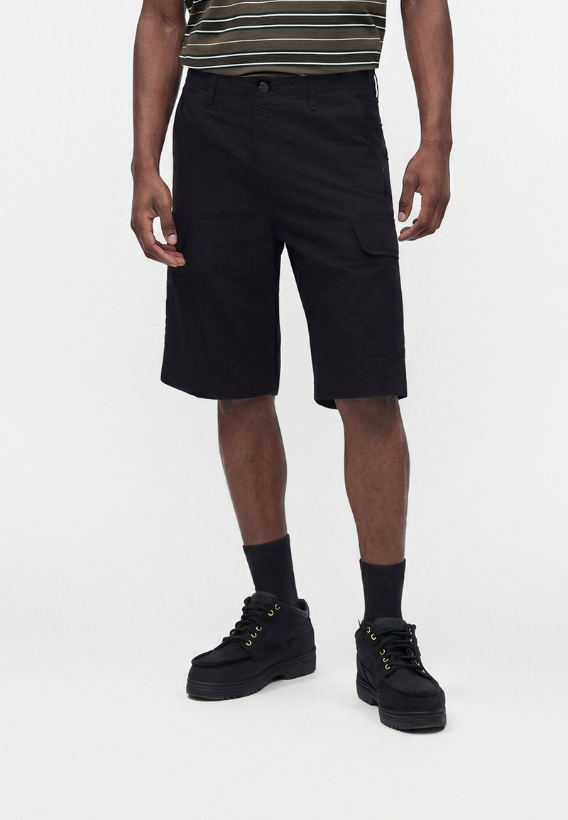 Homme portant un short cargo noir, des baskets montantes noires et des chaussettes noires, debout devant un fond clair uni.