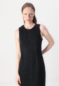 Black denim sleeveless dress featuring a round neckline, front seam detailing, and subtle side pockets. Smooth texture with structured shape.