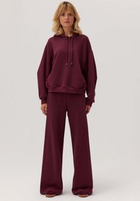 Bordowa bluza z kapturem i szerokie spodnie w zestawie z miękkiego materiału. Bluza posiada sznurki, a spodnie mają elastyczny pas i kieszenie.