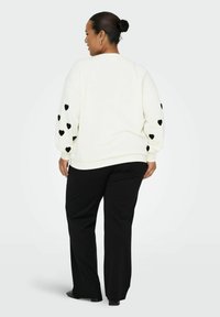 Sweatshirt blanc avec des motifs en cœur noirs sur les manches, associé à un pantalon noir. Le tissu semble doux avec une coupe décontractée.