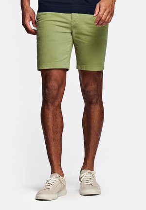 Man draagt lichtgroene korte broek en witte sneakers, staand tegen een eenvoudige witte achtergrond.