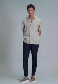 Lichtbeige shirt met korte mouwen, marineblauwe gestreepte tapered broek en grijze instappers, tegen een effen lichte achtergrond.