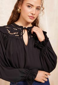 Blusa nera con colletto alto, design con chiave e dettagli in pizzo. Presenta maniche a sbuffo e polsini elasticizzati. Texture liscia e vestibilità fluida.