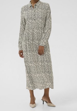 Femme portant une robe blanche à manches longues, longueur jusqu'aux chevilles, ornée de motifs abstraits noirs en forme de vagues, et des escarpins slingback beige à bout pointu.