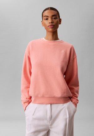 Kvinde iført en lyserød sweatshirt med et lille CK-logo på brystet og hvide bukser, stående med hænderne i lommerne mod en ensfarvet grå baggrund.