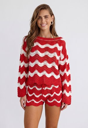 Vrouw glimlachend, gekleed in een rood-wit golvend gestreept gebreid top met lange mouwen en bijpassende korte broek tegen een effen achtergrond.