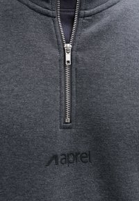 Nahaufnahme eines dunkelgrauen Stoffes mit teilweise geöffnetem silbernem Reißverschluss und darunter erhabenem schwarzem "aprel"-Logo.