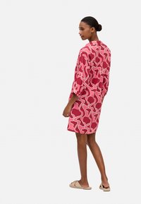 Vestido rojo con patrón ondulado rosa, mangas tres cuartos y un corte relajado, combinado con sandalias planas beige. Textura de tela suave.