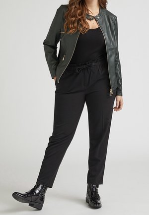 Femme portant une veste en cuir vert foncé, un haut noir, un pantalon noir à cordon et des bottines noires brillantes, debout avec un pied légèrement levé.