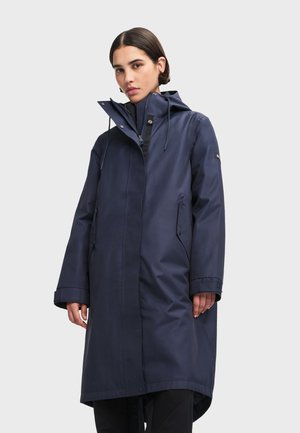 Femme portant un imperméable long bleu marine avec capuche, fermeture éclair frontale et poches à rabat latérales, debout devant un fond clair uni.