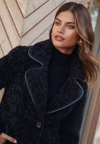 Palton negru, cu textură și aspect plușat, având margini negre lucioase și închidere cu nasturi. Purta peste un pulover înalt, cu guler și textură striată, de culoare închisă.