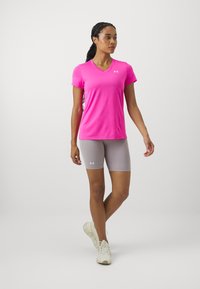 Ružičasta sportska majica kratkih rukava s V-izrezom, u kombinaciji s svijetlosivim kratkim hlačama. Oba komada imaju bijeli Under Armour logo.
