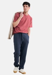 Pánsky outfit pozostávajúci z koralového bavlneného trička, námorníckych ľanových nohavíc a bielych tenisiek s béžovým saka prehodeným cez rameno.