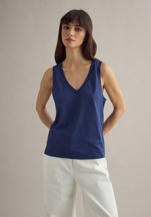 Donna con capelli lunghi e scuri che indossa una canotta blu navy con scollo a V e pantaloni bianchi, in piedi con le mani dietro la schiena.