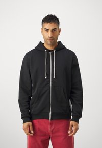 Sudadera con cremallera negra y capucha regulable, con bolsillos tipo canguro y puños acanalados, combinada con pantalones rojos. Textura de tela suave.