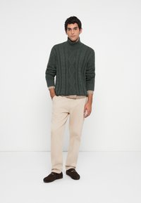 Homem de suéter de gola alta em ponto trançado verde escuro, calças beje claras, camisa xadrez à vista e sapatos loafers castanhos escuros, de pé contra uma parede branca.