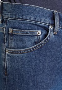 Nära bild av blå denimjeans som visar framfickan med nitar, bältesslinga och orange sömnadsdetaljer.