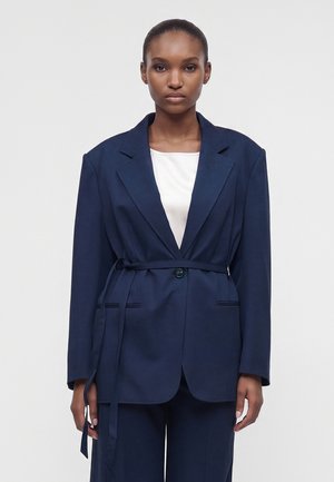 Femme à la peau foncée portant un blazer bleu marine ceinturé par-dessus un haut blanc, debout devant un fond clair uni.