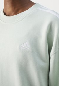 Mint green sweatshirt with ribbed collar. Features white Adidas logo and three white stripes on sleeves. Soft textured fabric.
