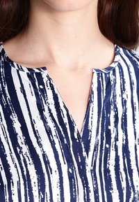 Blusa a righe blu navy e bianca con scollo a V e maniche corte, tessuto strutturato, caratterizzata da strisce verticali e un sottile dettaglio con bottoni.
