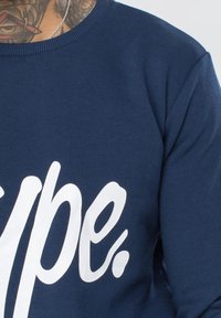 Sudadera azul marino con cuello acanalado, que presenta un gran texto en blanco que dice "hype". La tela parece suave y tiene una textura lisa.