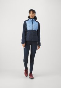 Blå og marineblå isolert jakke med glidelås i front, to sidelommer og hette, kombinert med marineblå leggings og røde joggesko.