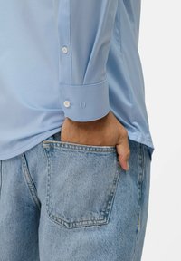 Hellblaues Langarmhemd mit geknöpften Ärmeln, kombiniert mit hellblauen Jeansshorts mit einer Gesäßtasche und lockerer Passform.
