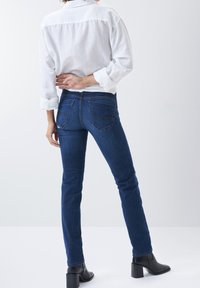 Persona vistiendo jeans azul oscuro y una camisa blanca abotonada, de pie con una mano detrás de la espalda contra un fondo sencillo.