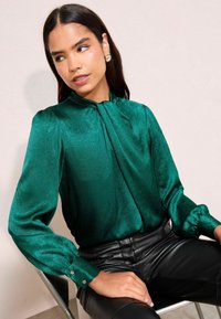 Roheline satiinkleit sidumisnööbiga ja puhvitud varrukatega, millel on nööpdetailid mansettides, kombineeritud mustade nahkpükstega.