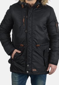 Veste rembourrée noire avec capuche en fausse fourrure, fermetures éclair et boutons, deux poches avant avec accents marron et poignets élastiques.