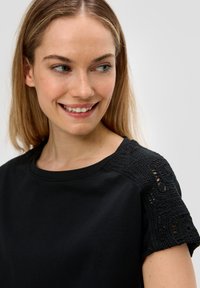 Czarny t-shirt z krótkim rękawem, ozdobiony skomplikowanym koronkowym detalem na ramionach, wykonany z miękkiego materiału i w klasycznym dekolcie w serek.