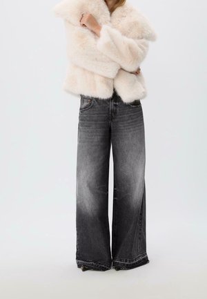 Personne portant une veste en fourrure duveteuse beige clair et un jean noir délavé taille haute à jambes larges, debout devant un fond uni.