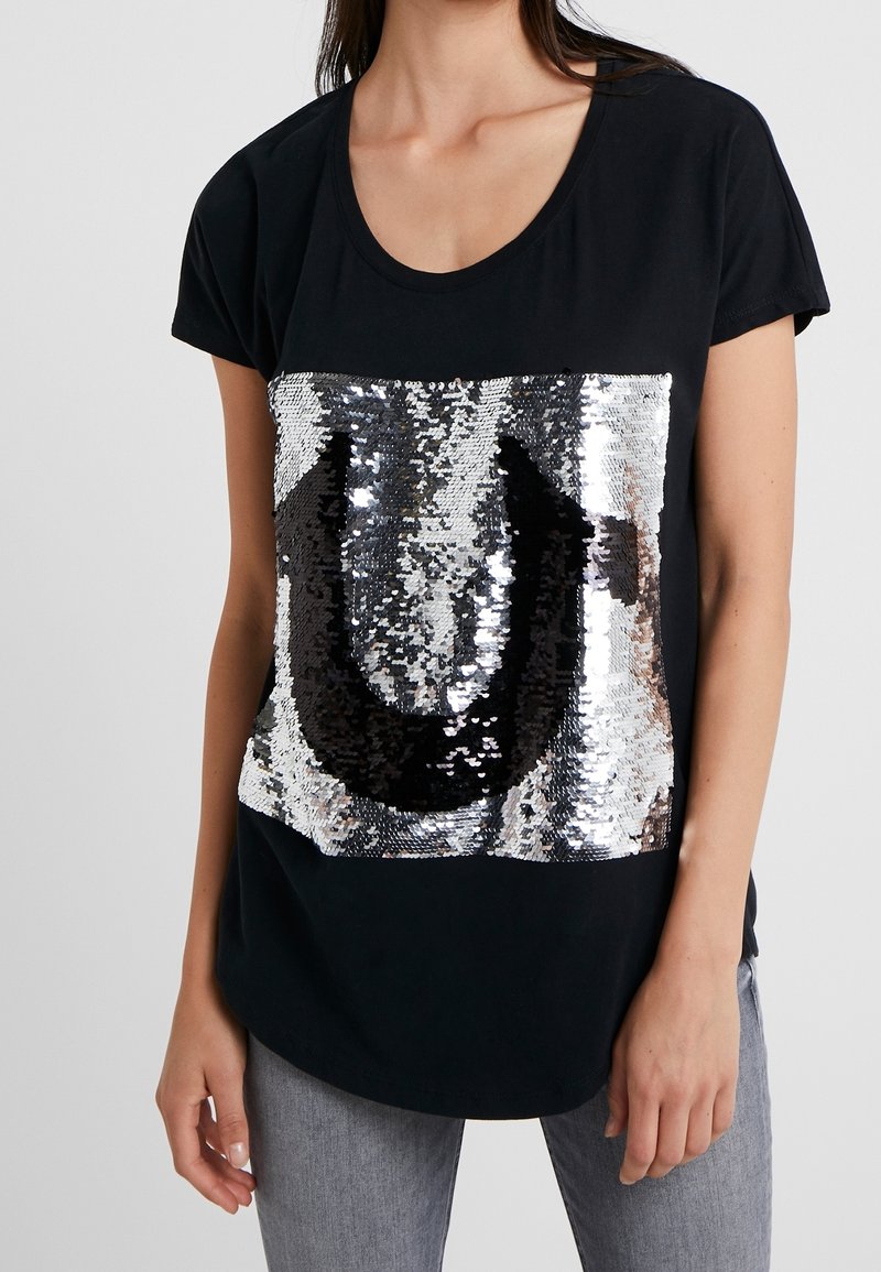 Svart T-shirt med en stor, paljettprydd design i silver och svart som liknar bokstaven "U". Kort ärm och rundad halsringning.