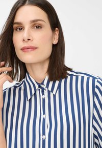 Camicia a righe blu e bianche con colletto appuntito, chiusura anteriore con bottoni e texture del tessuto liscia. Capelli parzialmente pettinati di lato, che mostrano un orecchino semplice.