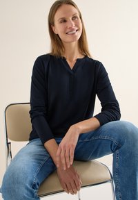 Haut à manches longues bleu marine avec un col en V et une coupe décontractée, associé à un jean bleu clair, assis sur une chaise minimaliste. Texture de tissu lisse.