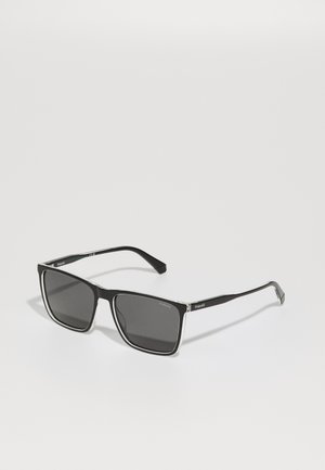 Lunettes de soleil rectangulaires noires avec des verres teintés gris, présentant un fin accent en argent le long de la monture, et un design en plastique élégant.