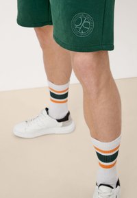 Shorts en coton verts avec un logo imprimé, assortis à des baskets blanches avec des accents noirs, et des chaussettes blanches avec des rayures orange et vertes.