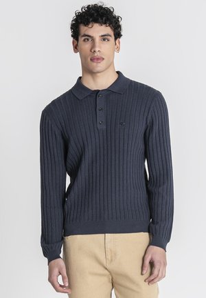 Hombre con cabello rizado que lleva un jersey polo de manga larga acanalado de color gris oscuro y pantalones beige, de pie frente a un fondo liso.